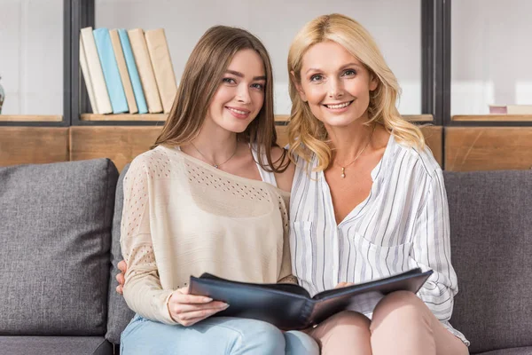 Glückliche Mutter und Tochter lächeln in die Kamera, während sie auf dem Sofa sitzen und ein Fotoalbum in der Hand halten — Stockfoto