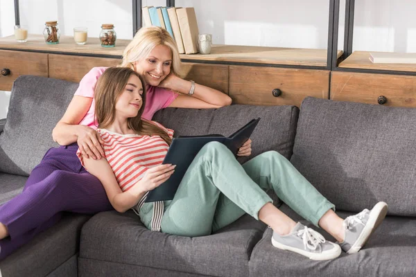 Щаслива жінка з молодою дочкою відпочиває на дивані і дивиться на фотоальбом — стокове фото
