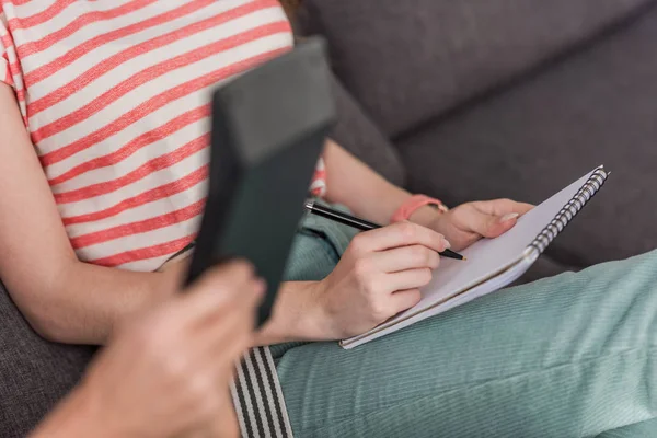 Vista recortada de la niña escribiendo en un cuaderno cerca de la madre que sostiene la calculadora - foto de stock