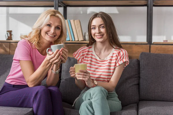 Mãe alegre e filha olhando para a câmera enquanto sentado no sofá e segurando xícaras de chá — Fotografia de Stock