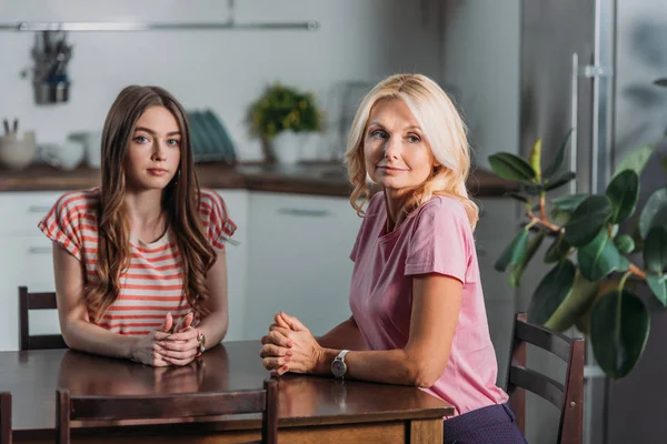 Chica bonita mirando a la cámara mientras está sentado en la mesa de la cocina cerca de la madre - foto de stock