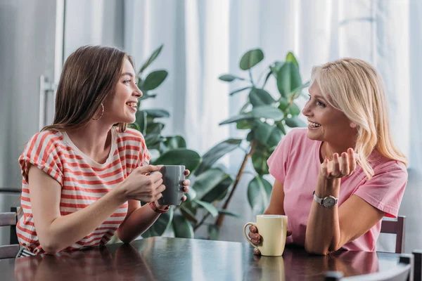 Alegre hija y madre sentado en la mesa de la cocina, hablando y bebiendo té - foto de stock