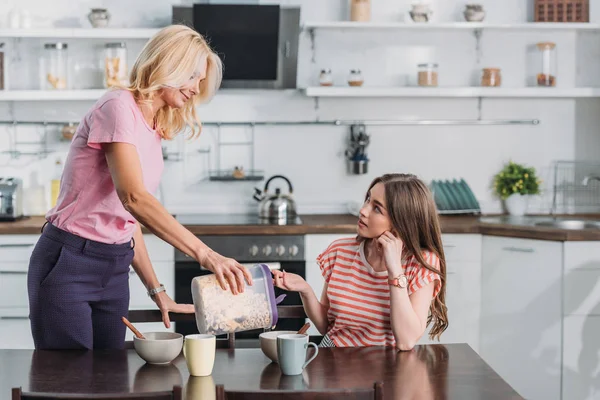 Attraktive reife Frau fügt Flocken in Schüssel hinzu, während ihre kleine Tochter am Küchentisch sitzt — Stockfoto