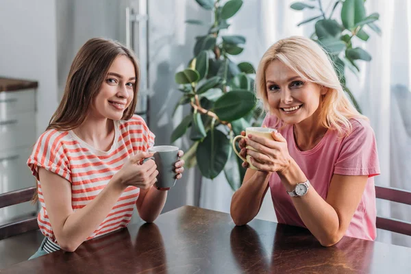 Heureux mère et fille souriant à la caméra tout en étant assis à la table de cuisine avec des tasses à thé — Photo de stock