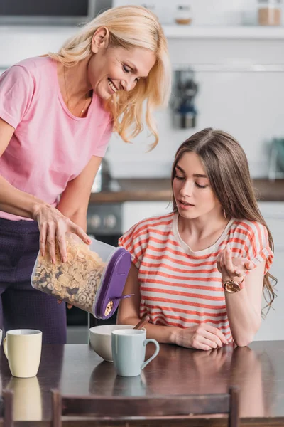 Lächelnde reife Frau fügt Flocken in Schüssel hinzu, während ihre kleine Tochter am Küchentisch sitzt — Stockfoto