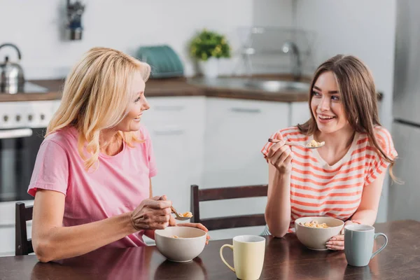 Feliz madre e hija hablando mientras están sentadas en la mesa de la cocina y desayunando - foto de stock