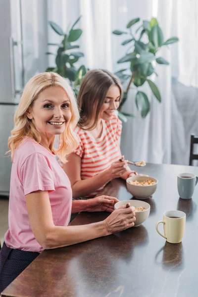 Heureux mature femme regardant caméra tout en prenant le petit déjeuner avec jeune fille — Photo de stock