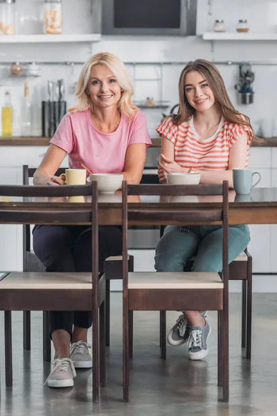 Lächelnde Mutter und Tochter blicken in die Kamera, während sie am Küchentisch sitzen und frühstücken — Stockfoto