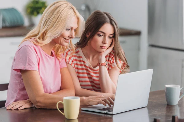 Attentive girl sitting at kitchen table near smiling mother using laptop — Stock Photo