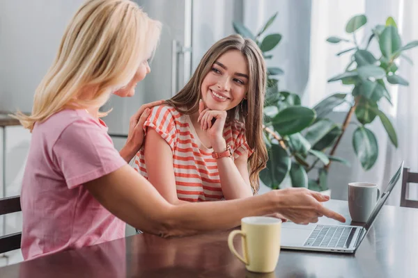 Mutter zeigt auf Laptop, während sie die Schulter ihrer kleinen, lächelnden Tochter berührt — Stockfoto