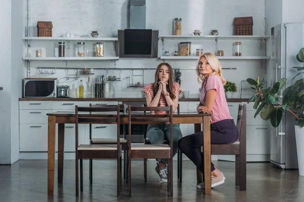 Attractive mature woman with young girl sitting at kitchen table and looking at camera — Stock Photo