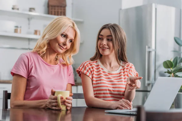 Ragazza allegra che punta con il dito al computer portatile mentre si siede al tavolo della cucina vicino alla madre sorridente — Foto stock