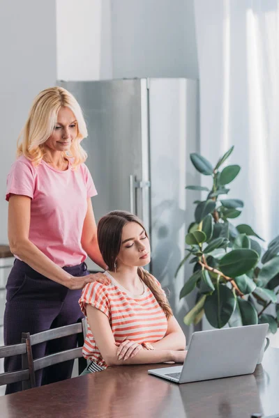 Beautiful mature woman touching shoulders of young daughter sitting near laptop in kitchen — Stock Photo