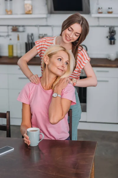 Cheerful girl touching sholuders of mother sitting at kitchen table with tea cup — Stock Photo