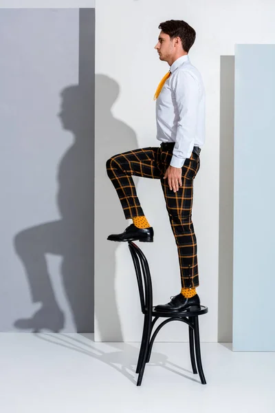 Vista lateral del hombre en pantalones a cuadros de pie en silla en blanco y gris - foto de stock
