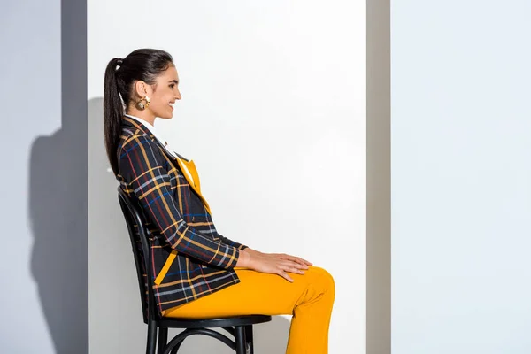 Seitenansicht einer fröhlichen jungen Frau, die auf einem Stuhl in grau und weiß sitzt — Stockfoto