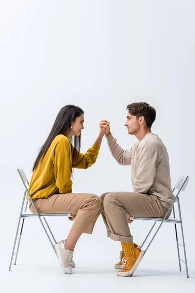 Side view of handsome man and woman holding hands while sitting on chairs on white — Stock Photo