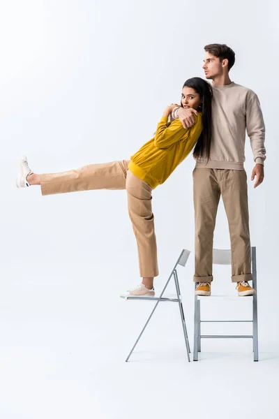 Schöner Mann steht auf Stuhl und umarmt stilvolles Mädchen auf weißem Grund — Stockfoto