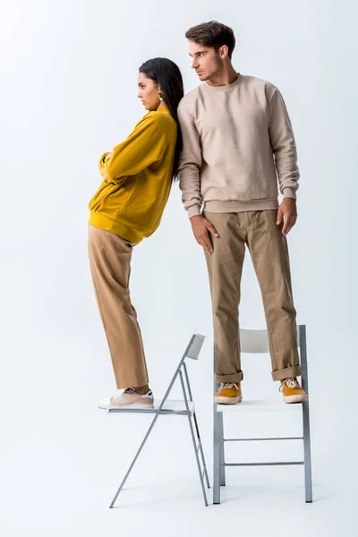 Стильная девушка позирует рядом с мужчиной, стоящим на стуле на белом — стоковое фото