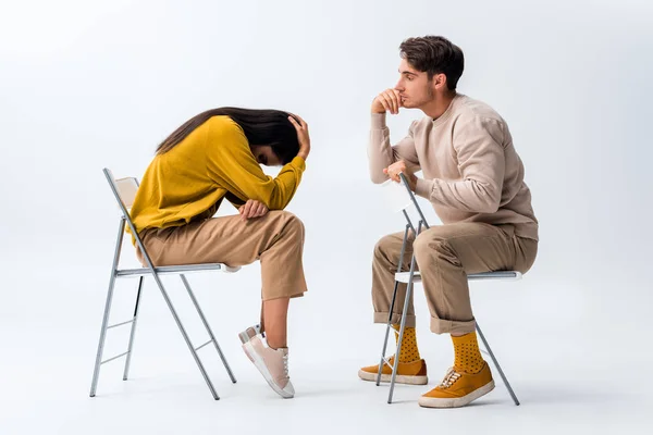 Hombre sentado en la silla y mirando a la mujer molesta cubriendo la cara en blanco - foto de stock