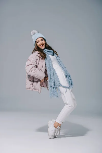 Attraente ragazza sorridente che balla in cappotto caldo, cappello e sciarpa in maglia su grigio — Foto stock