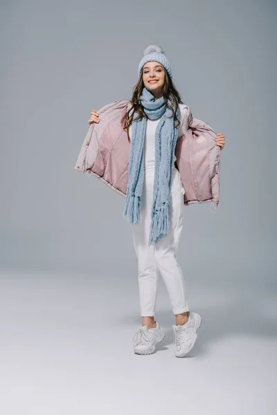 Happy girl posing in warm coat, knitted hat and scarf on grey — Stock Photo