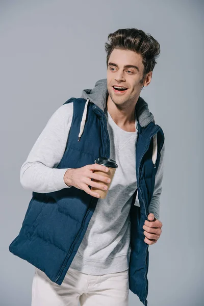 Веселий чоловік у теплому жилеті тримає каву, щоб піти, ізольований на сірому — стокове фото