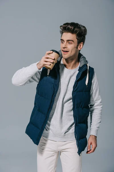 Happy handsome man in warm waistcoat drinking coffee to go, isolated on grey — Stock Photo