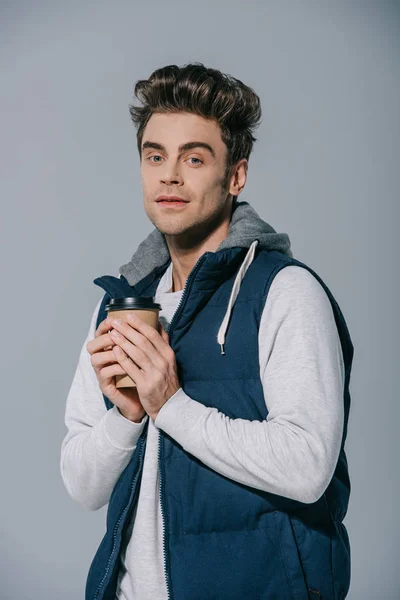 Hombre guapo en chaleco caliente sosteniendo café para llevar, aislado en gris - foto de stock