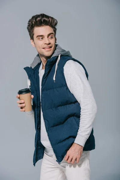 Styling homem sorridente em colete quente segurando café para ir, isolado em cinza — Fotografia de Stock