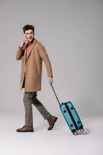 Lächelnder Mann im beigen Mantel, der mit Smartphone spricht und mit Reisetasche auf grau läuft — Stockfoto