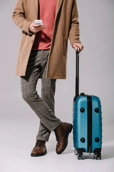 Vista recortada de hombre elegante de pie con bolsa de viaje y teléfono inteligente en gris - foto de stock