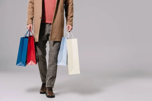Vista recortada de hombre con estilo en abrigo beige sosteniendo bolsas de compras en gris - foto de stock