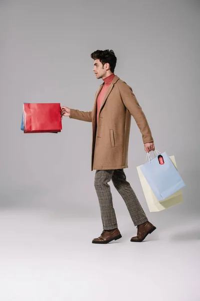 Side view of stylish man in beige coat walking with shopping bags on grey — Stock Photo
