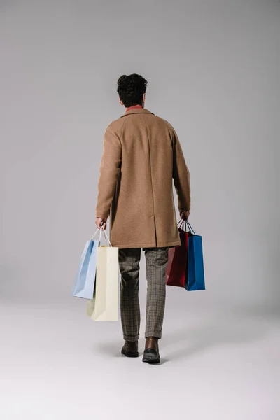 Vista trasera del hombre en abrigo beige sosteniendo bolsas de compras en gris - foto de stock