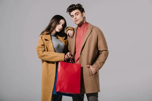 Casal na moda em casacos bege posando com sacos de compras isolados em cinza — Fotografia de Stock