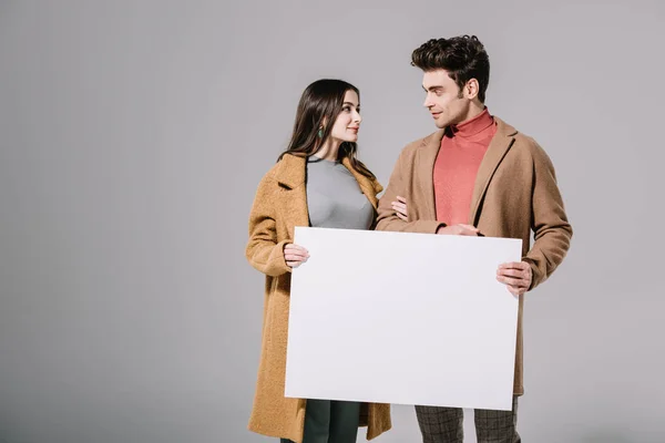 Beau couple élégant en manteaux beige posant avec planche vide, isolé sur gris — Photo de stock