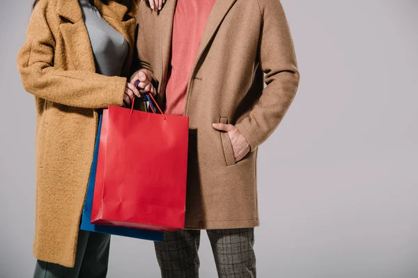 Vista recortada de pareja en abrigos beige sosteniendo bolsas aisladas en gris - foto de stock