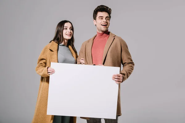 Cheerful couple in beige coats posing with empty board, isolated on grey — Stock Photo