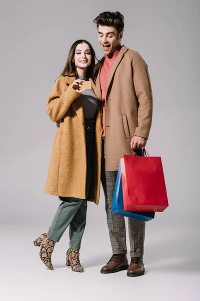 Happy couple in beige coats posing with shopping bags and credit card on grey — Stock Photo