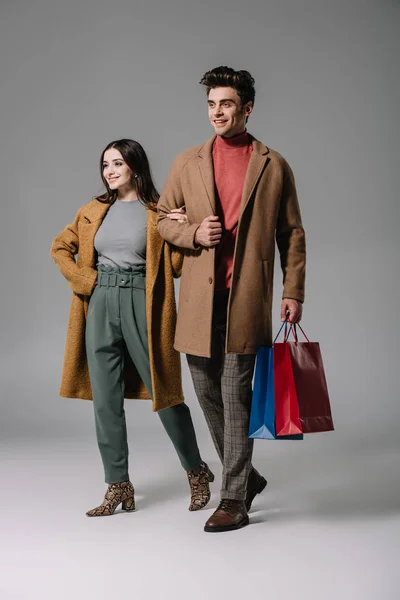 Pareja sonriente en abrigos beige posando con bolsas de compras en gris - foto de stock