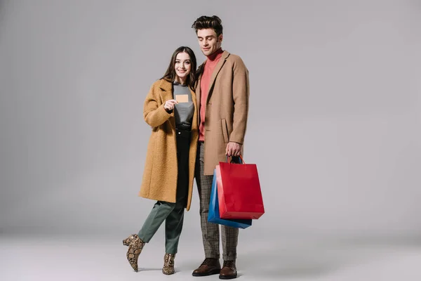 Beautiful couple in beige coats posing with shopping bags and credit card on grey — Stock Photo