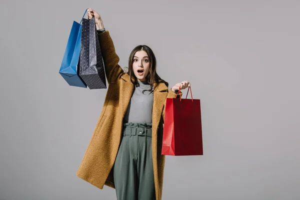 Belle fille choquée en manteau beige tenant des sacs à provisions, isolé sur gris — Photo de stock