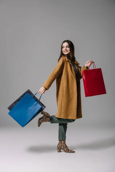 Модная улыбающаяся девушка в бежевом пальто ходит с пакетами для покупок на сером — стоковое фото