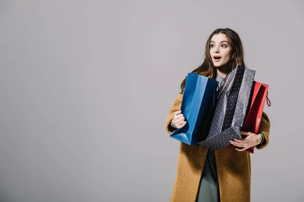 Attraente ragazza sorpresa in beige cappotto che tiene borse della spesa, isolato su grigio — Foto stock