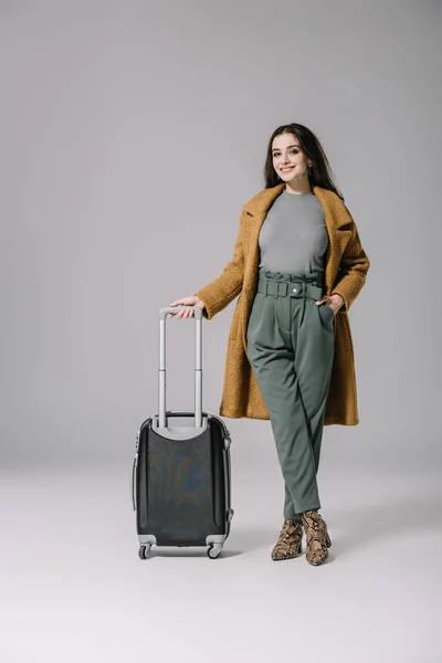 Beautiful smiling girl in beige coat posing with travel bag on grey — Stock Photo