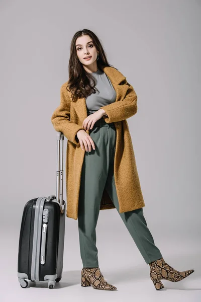 Hermosa mujer en abrigo beige posando con bolsa de viaje en gris - foto de stock