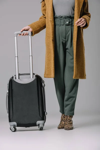 Vista ritagliata di donna elegante in cappotto beige in posa con borsa da viaggio su grigio — Foto stock