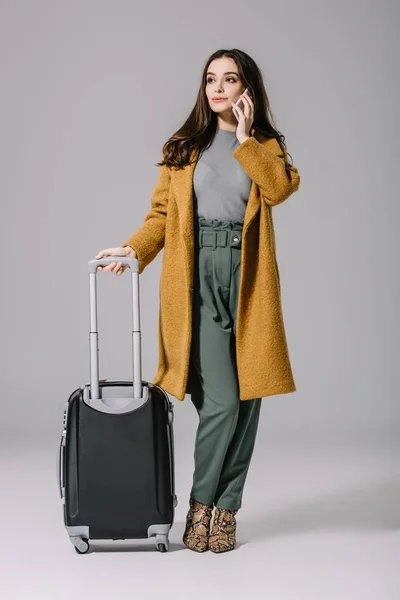 Красивая стильная девушка в бежевом пальто стоит с дорожной сумкой, разговаривая по смартфону на сером — стоковое фото