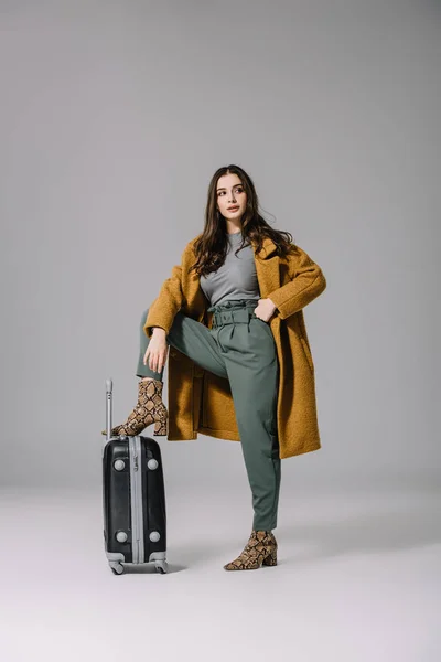 Hermosa mujer elegante en abrigo beige posando con bolsa de viaje en gris - foto de stock